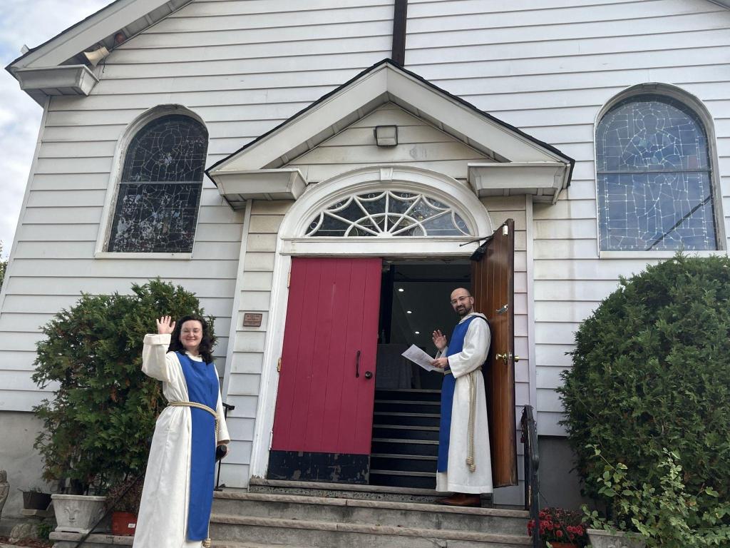 Two members of the Society in blue scapulars wave from the open red doors of the Church of the Atonement.